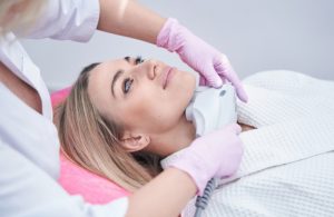 A female clinician in pink gloves performs a FaceTite procedure on a woman's jaw and neck area using a specialised handheld device in a clean, clinical setting.